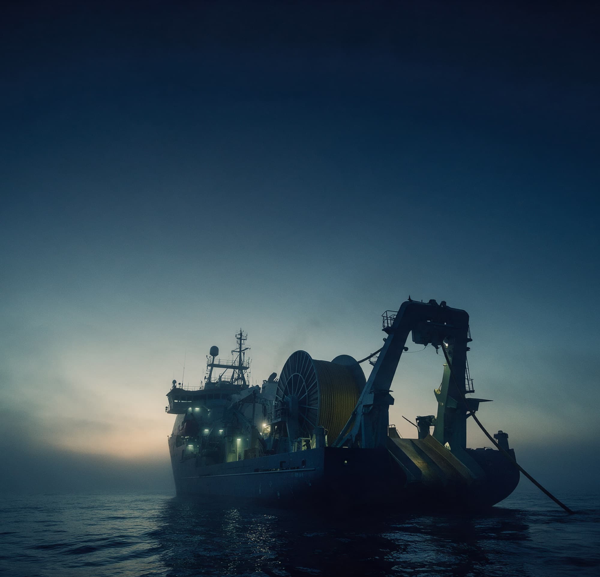 A cable-laying ship at sea at dawn, its cable drum and stern chute paying cable into the water